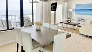 Una sala de estar moderna con comedor, ventanas amplias con vista al océano, un televisor de pantalla plana y un cuadro en la pared.