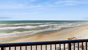 Vista a la playa desde un balcón con barandilla, olas del océano y un vehículo estacionado.
