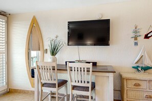 A room with a TV mounted on the wall, two white chairs, a wooden desk, and a decorative mirror. - Beachfront Serenity with Balcony, Walk to Dining & Attractions (Daytona Beach Shores)