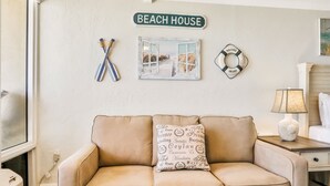 Ein beiges Sofa mit einem gemusterten Kissen, ein an der Wand befestigtes Schild mit der Aufschrift "BEACH HOUSE", ein Paar verschränkter Schläger, ein gerahmtes Bild von einer Strandansicht und ein Schild mit der Aufschrift "WELCOME ABOARD".