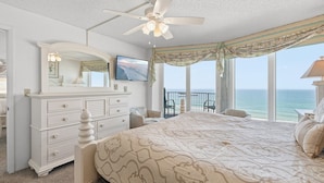 A bedroom with a large bed, a dresser, a ceiling fan, a TV, and a balcony with a view of the ocean. - Elegant Oceanfront Condo w/ Balcony, Pool & Steps to Beach (Daytona Beach Shores)