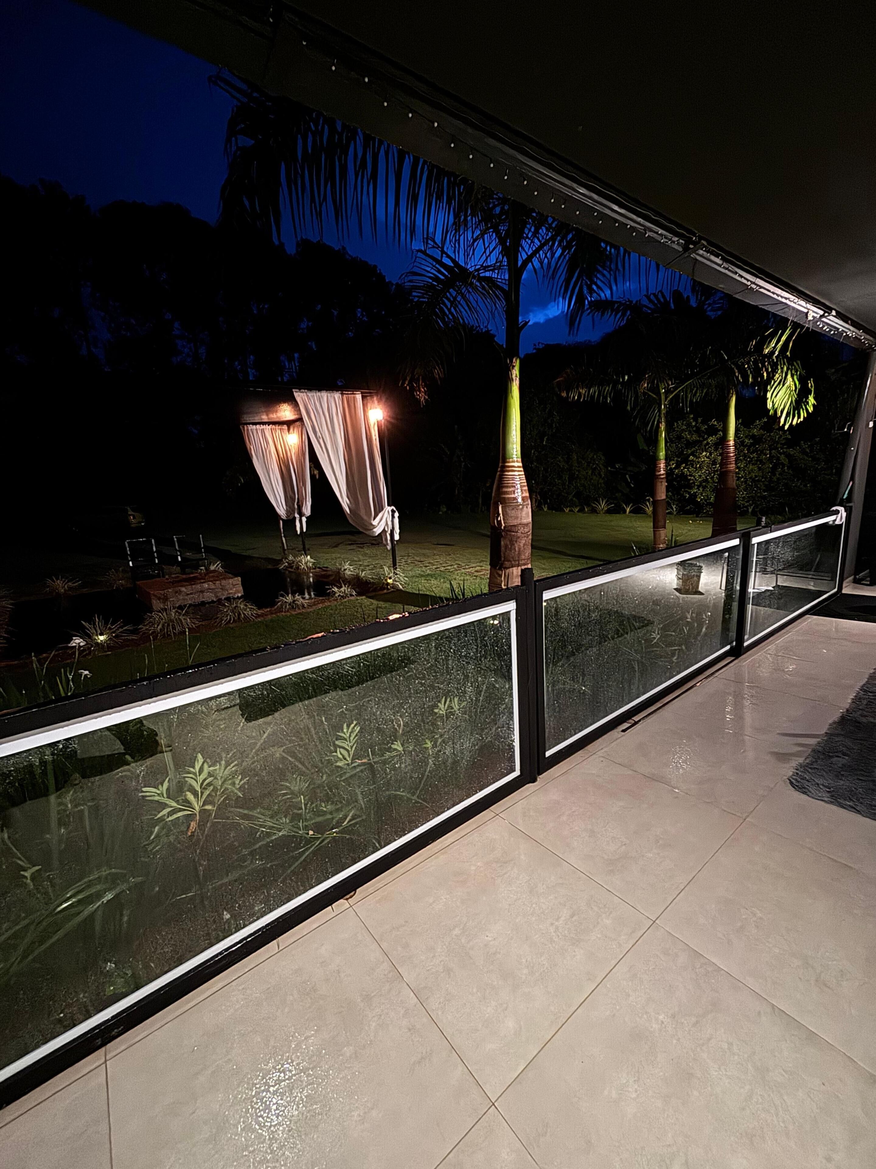 Un espace extérieur moderne doté d’une balustrade en verre, d’un coin salon avec une table et des chaises, et d’un décor composé de palmiers et d’un jardin.