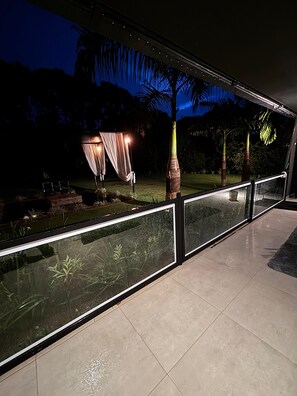 Een modern buitengebied met een glazen balustrade, een zithoek met een tafel en stoelen, en een achtergrond van palmbomen en een tuin.