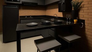 A modern kitchen with dark cabinetry, a microwave, a stove, and a sink. There are two stools at the counter and a potted plant on the wall. - Cozy House "exclusively for Couples" Pet Friendly (Bragança Paulista)
