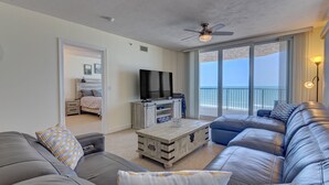 Een moderne woonkamer met een grijze leren bank, een houten salontafel en een televisie. Vanuit grote ramen is een balkon met uitzicht op de oceaan te zien.