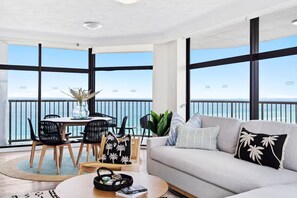 Ein modernes Wohnzimmer mit Balkon, großen Fenstern, einer Couch, einem Couchtisch und einer Essecke mit Stühlen und Tisch.