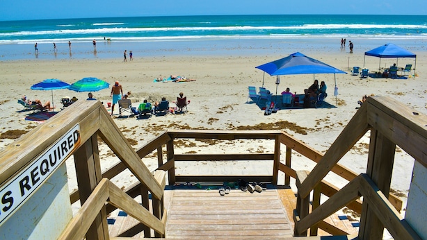 A wooden staircase leading to a beach with blue umbrellas and beach chairs. - Modern Coastal Condo w/ Balcony, Pool & Easy Walk to the Beach (New Smyrna Beach)