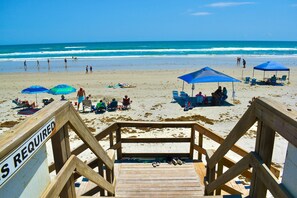 Eine Holztreppe führt zu einem Strand mit blauen Sonnenschirmen und Liegestühlen.