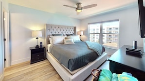 A bedroom with a bed, a nightstand, a desk, a ceiling fan, and a window with a view of buildings. - Modern Coastal Condo w/ Balcony, Pool & Easy Walk to the Beach (New Smyrna Beach)