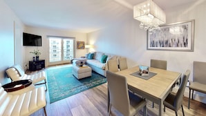 A modern living room with a sofa, a dining table, and a TV mounted on the wall. - Modern Coastal Condo w/ Balcony, Pool & Easy Walk to the Beach (New Smyrna Beach)