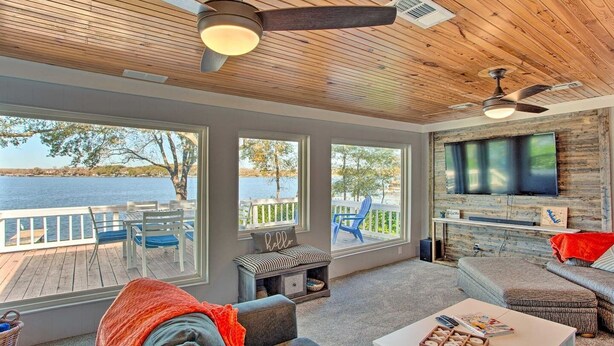 A spacious room with a large window overlooking a body of water, a ceiling fan, a television mounted on a wooden wall, and a cozy seating area with a bench and a sofa. - Dock & Fire Pit: Lakefront Cedar Creek Home! (Mabank)