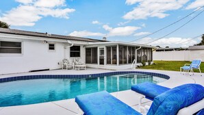 A swimming pool with blue and white lounge chairs, a covered patio, and a white house with a screened porch. - Quiet 4BR Getaway w/Private Pool, Walk to Beach & Florida Room (Ormond Beach)