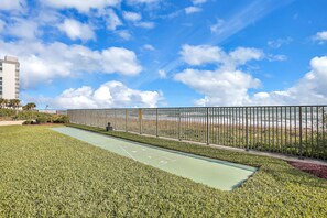 A green shuffleboard court with a metal fence and a view of the ocean. - Oceanfront escape w/ 2 balconies & pool access – peaceful beach vibes (Ormond Beach)