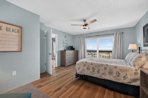 A bedroom with a bed, a ceiling fan, a dresser, a window with curtains, and a view of the beach. - Oceanfront escape w/ 2 balconies & pool access – peaceful beach vibes (Ormond Beach)