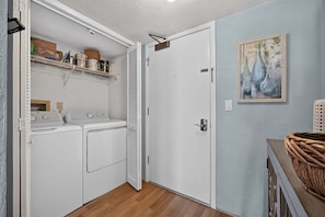 A laundry room with a washing machine and dryer, a shelf with cleaning supplies, and a door leading to another room. - Oceanfront escape w/ 2 balconies & pool access – peaceful beach vibes (Ormond Beach)