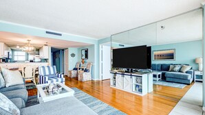 Una sala moderna con sofá, sillones, una mesita de centro, televisor y una cocina al fondo.