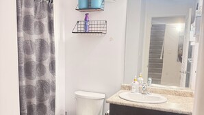 Ein Badezimmer mit Toilette, Waschbecken und Spiegel. Zwei Pflanzen stehen auf Regalen, ein Duschvorhang ist angebracht und ein Teppich liegt auf dem Boden.