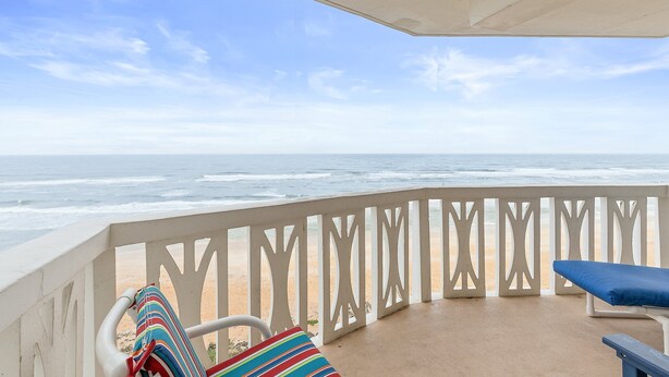 A balcony with a view of the ocean, a white railing, and a blue cushioned chair.