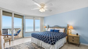 A bedroom with a large bed, a nightstand, a chair, and a view of the ocean through a sliding door.