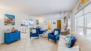 Un salon moderne avec un canapé beige, des fauteuils bleus et une commode bleue. Les murs sont ornés de tableaux et une grande fenêtre laisse entrer la lumière naturelle.