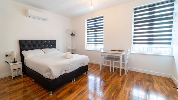 A bedroom with a bed, bedside table, lamp, and a desk with a chair. - Centra High Road (London)