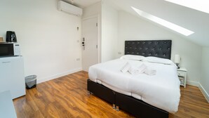 A bedroom with a bed, a bedside table, a lamp, a microwave, and a trash bin. - Centra High Road (London)