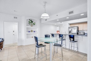 A modern kitchen with a glass dining table, four chairs, and a hanging light fixture. - Oceanwalk 3BR Condo w/ Pool, Tennis & Balcony Across from Beach (New Smyrna Beach)