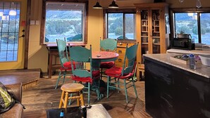 Een gezellig cabin interieur met houten plafond, een eethoek met verschillende stoelen, een keukenhoek en grote ramen met uitzicht op een besneeerd landschap.