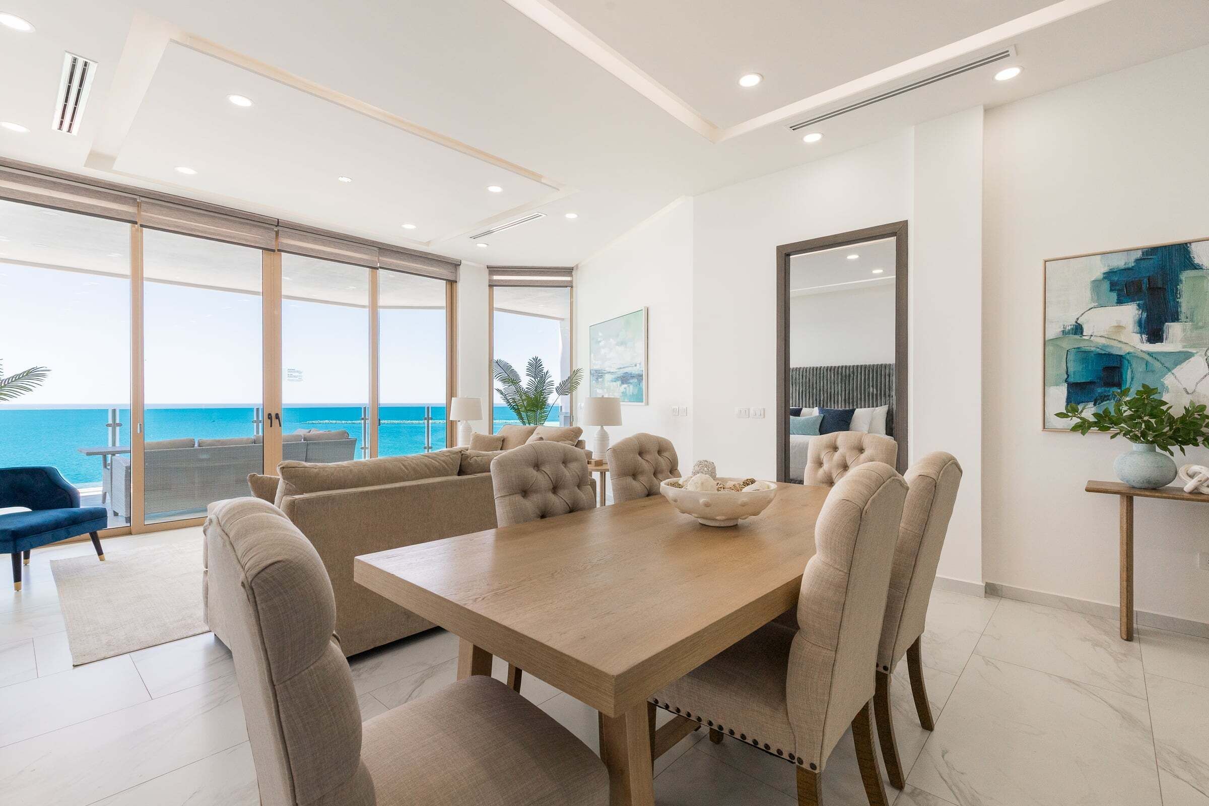 A modern dining area with a wooden table, beige upholstered chairs, and a view of the ocean.