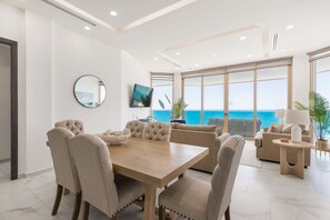 Un salon moderne avec une table à manger en bois, des chaises recouvertes d’un tissu beige, un grand téléviseur à écran plat et une vue sur l’océan.