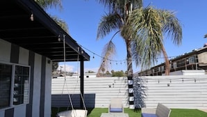 A patio with outdoor furniture, including a swing chair and a table, surrounded by artificial grass and palm trees. - Exotic home 3bed2bath 20min to beach downtown (La Presa)