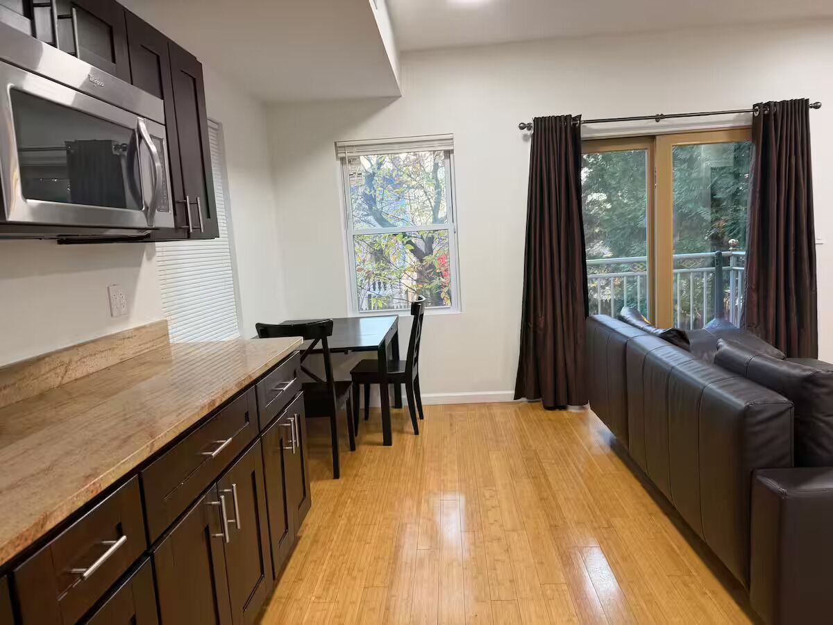 A kitchen with a microwave, cabinets, and a dining table with chairs. There is a window with curtains and a view of trees.