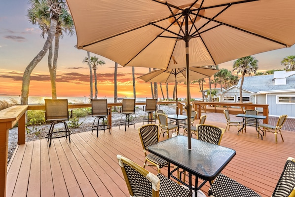 A wooden deck with outdoor seating, including tables and chairs, overlooking a beach with palm trees and a sunset.
