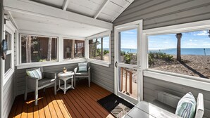 En inglasad veranda med trägolv, vita väggar och stora fönster som ger utsikt över stranden.
