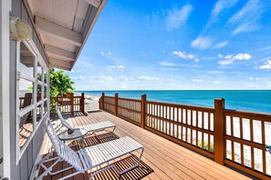 Eine Holzterrasse mit weißen Metallstühlen und einem Tisch, mit Blick auf einen Strand mit Holzzaun.