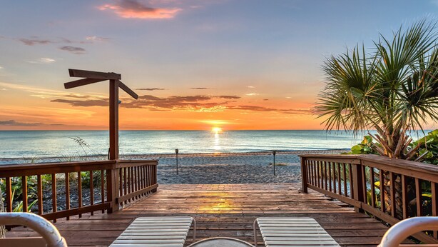 Een houten terras met loungebedden, gericht op het strand bij zonsondergang.
