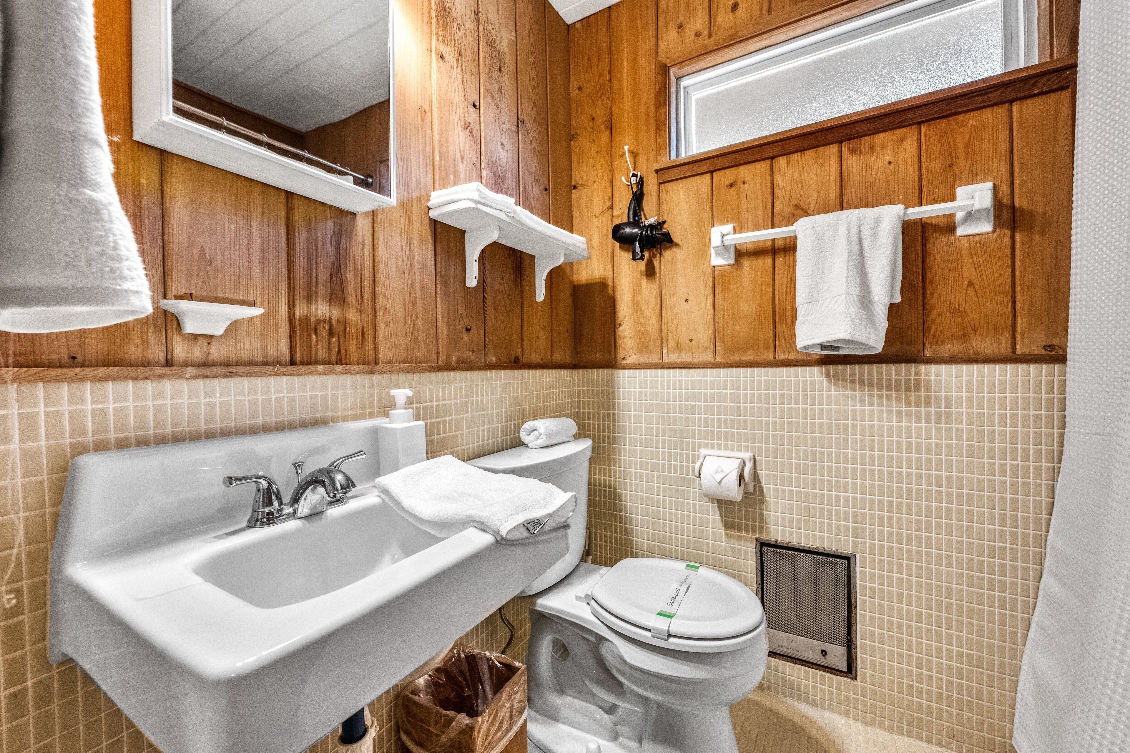 A bathroom with wooden paneling, a white sink, a toilet, and a window above the mirror.