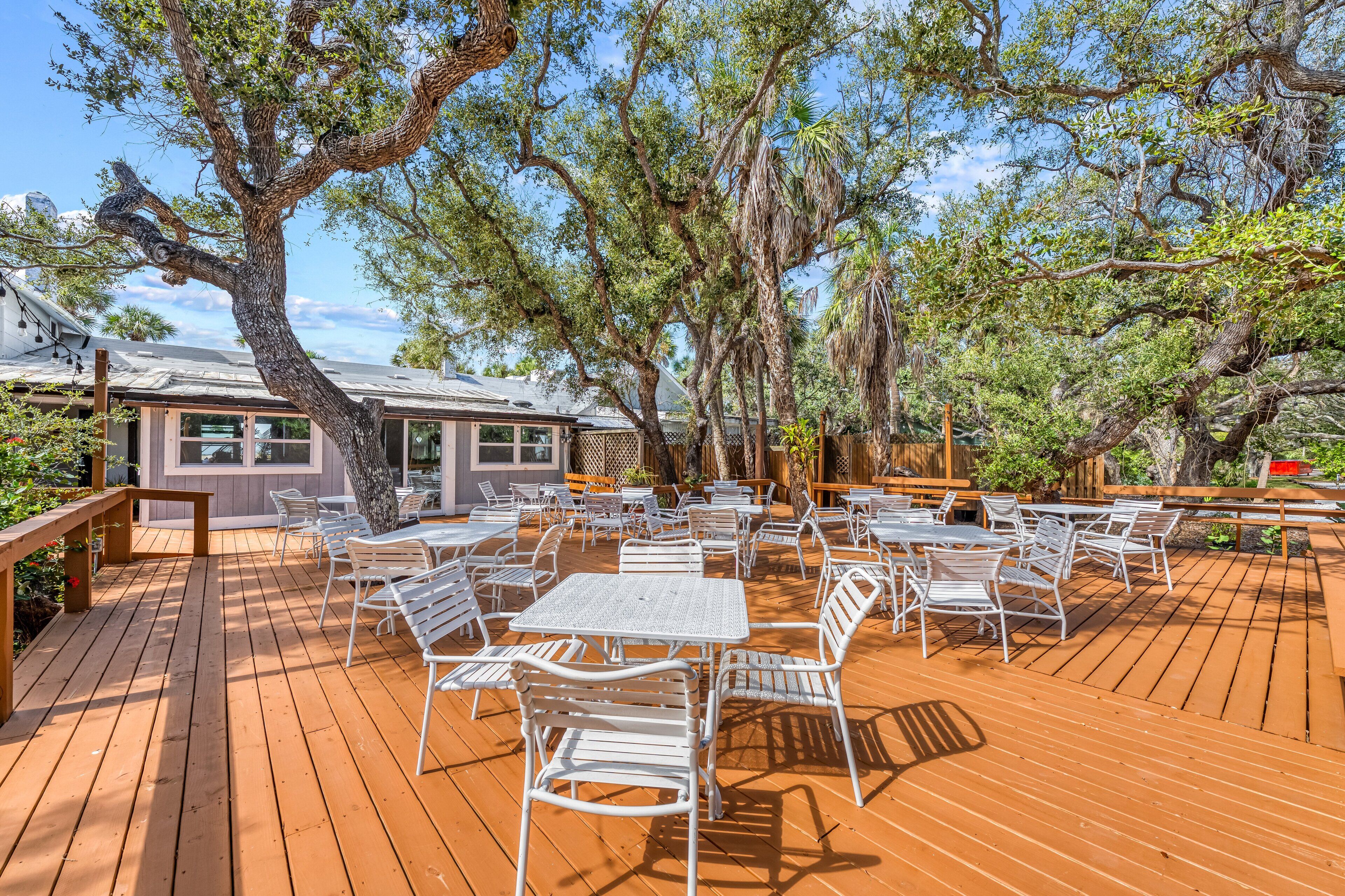 Une terrasse extérieure avec des tables et des chaises en métal blanc, entourée d’arbres et d’une plateforme en bois.