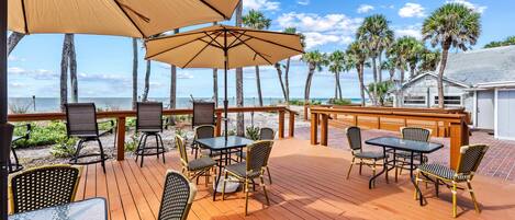 A wooden deck with outdoor seating, including tables and chairs, overlooking a beach with palm trees and a clear sky.