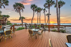A wooden deck with outdoor seating, overlooking a beach with palm trees and a sunset. - Gulf Front Escape on the Beach with Shared Viewing Deck & Pool - Cozy & Intimate (Englewood)