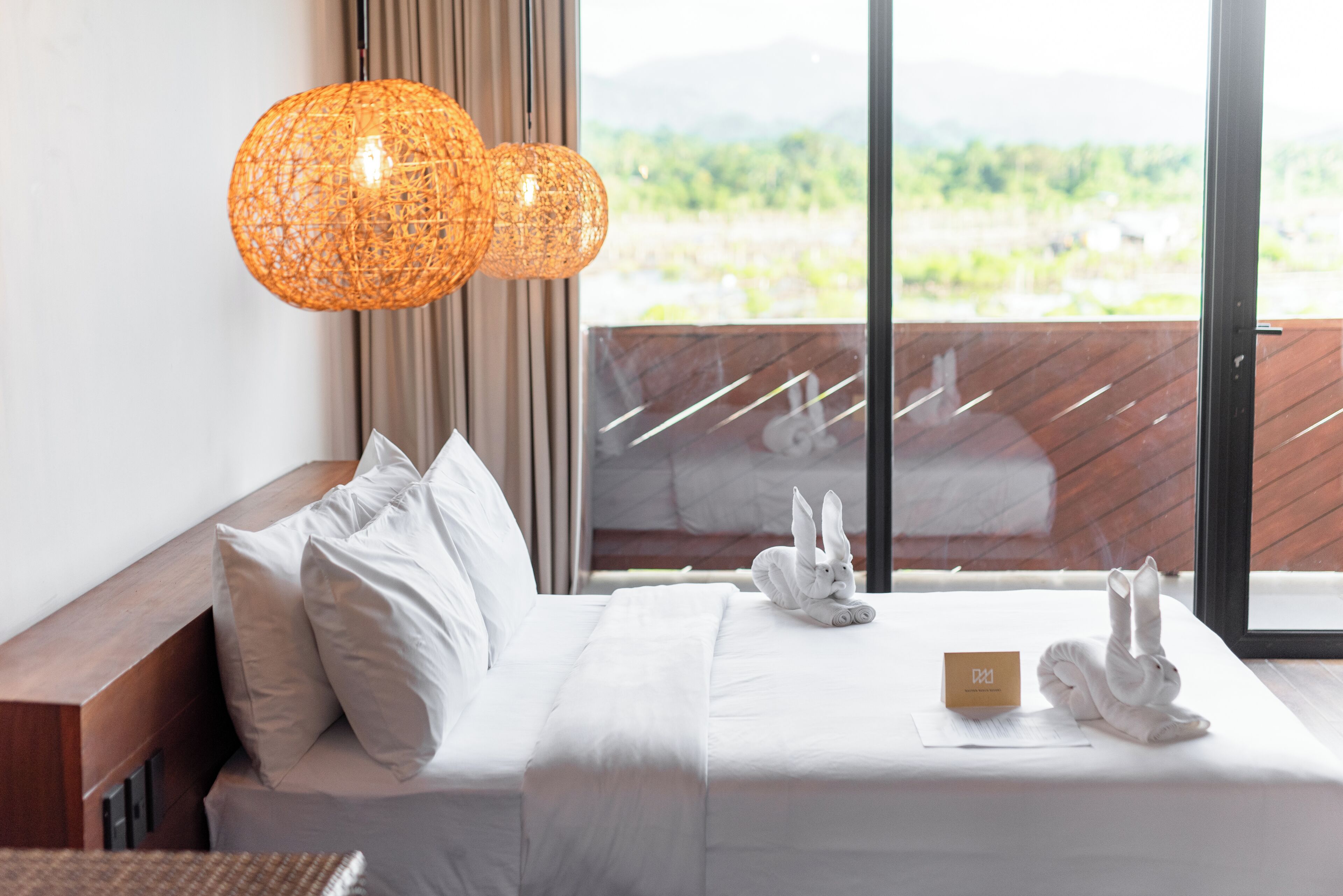 A hotel room with a bed, white bedding, a name card, and a view of greenery through the window.