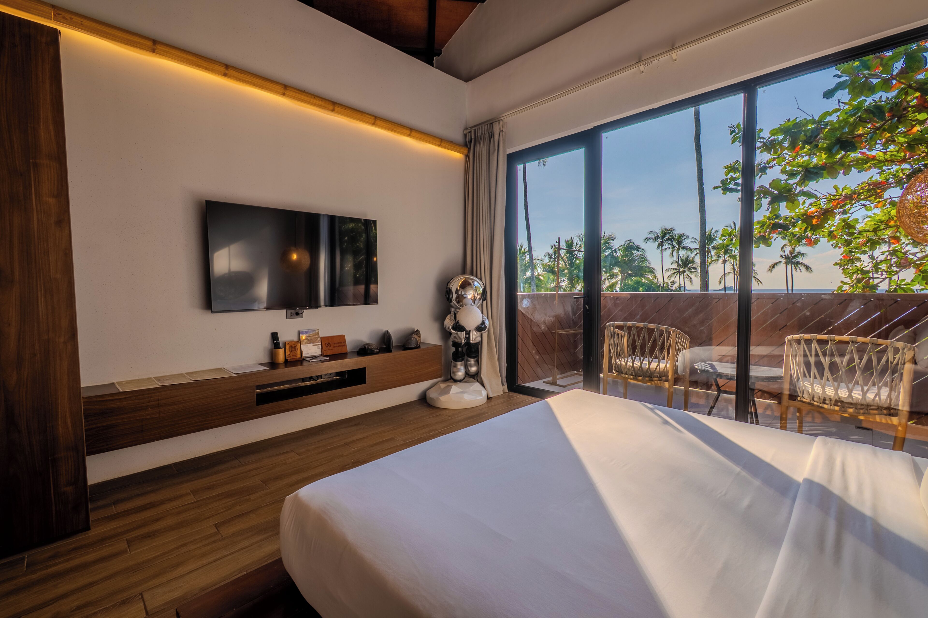 A modern hotel room with a large bed, a TV mounted on the wall, a wooden bedside table, and a balcony with wicker chairs.