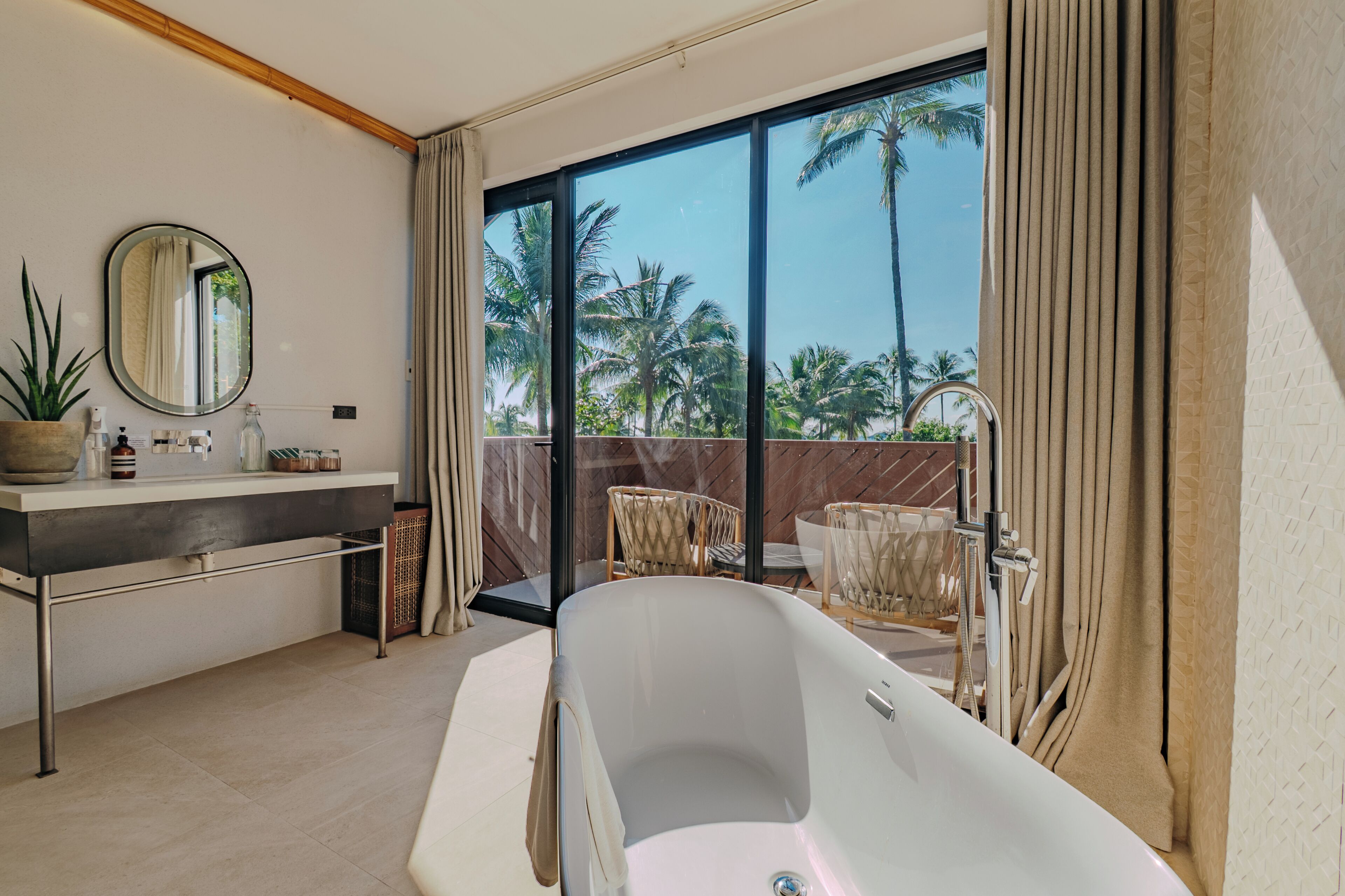 Ein modernes Badezimmer mit einer Badewanne, einem Waschbecken mit Spiegel und einem großen Fenster mit Blick auf Palmen.