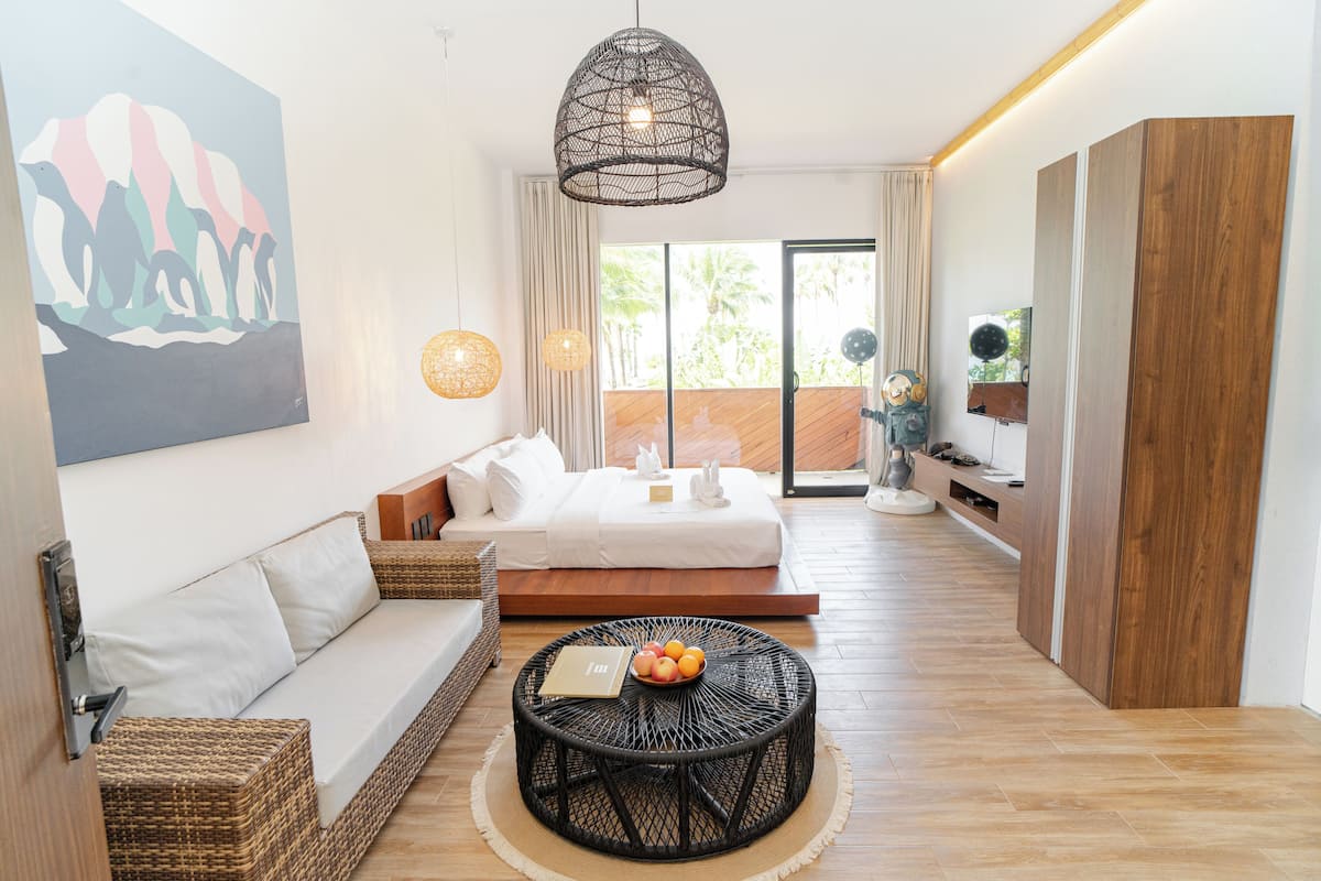 Une chambre d’hôtel moderne avec un lit, un canapé, une table ronde avec des fruits, des œuvres d’art et une grande fenêtre.