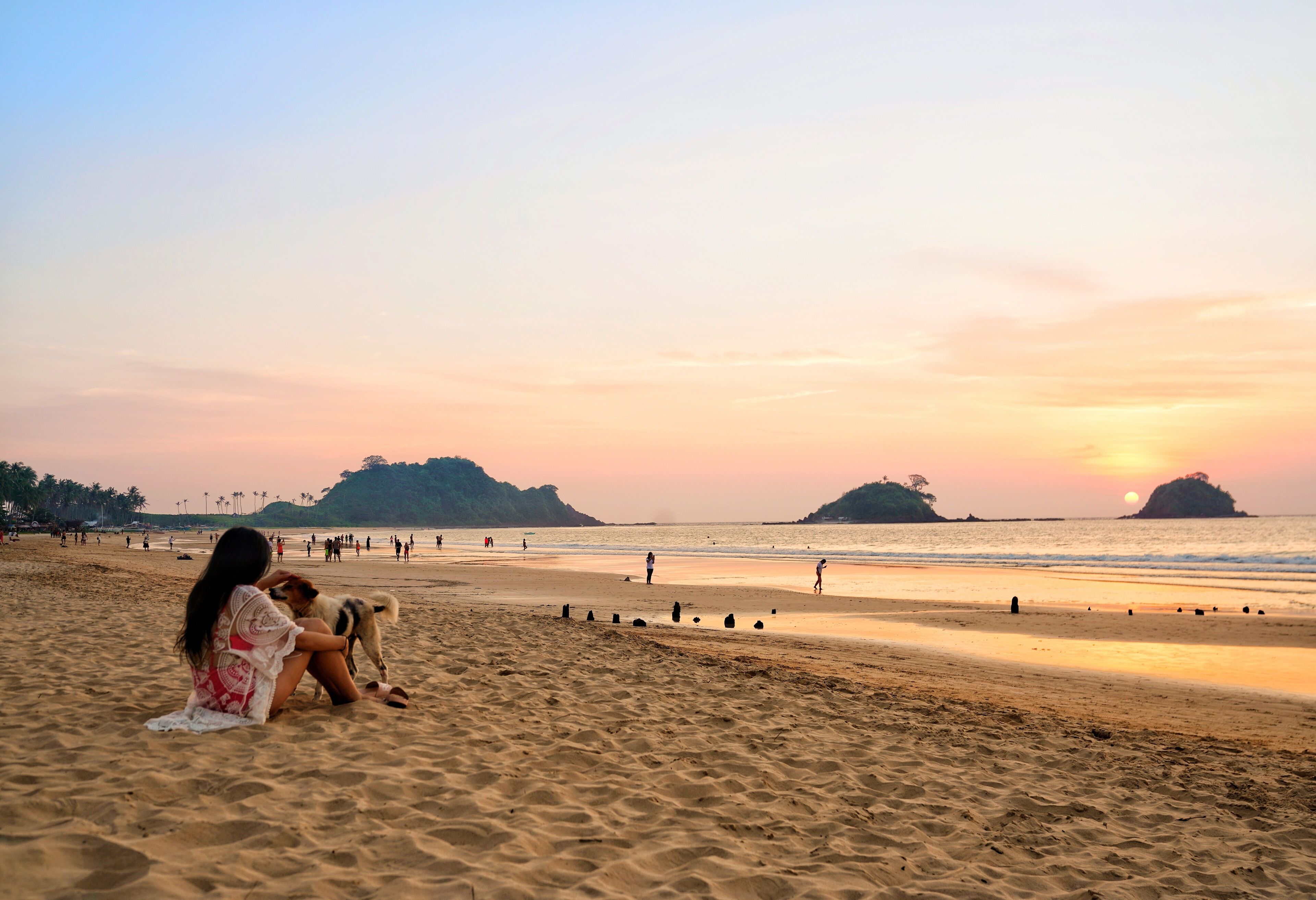 A beach scene with people and a dog, featuring a sunset and distant islands.