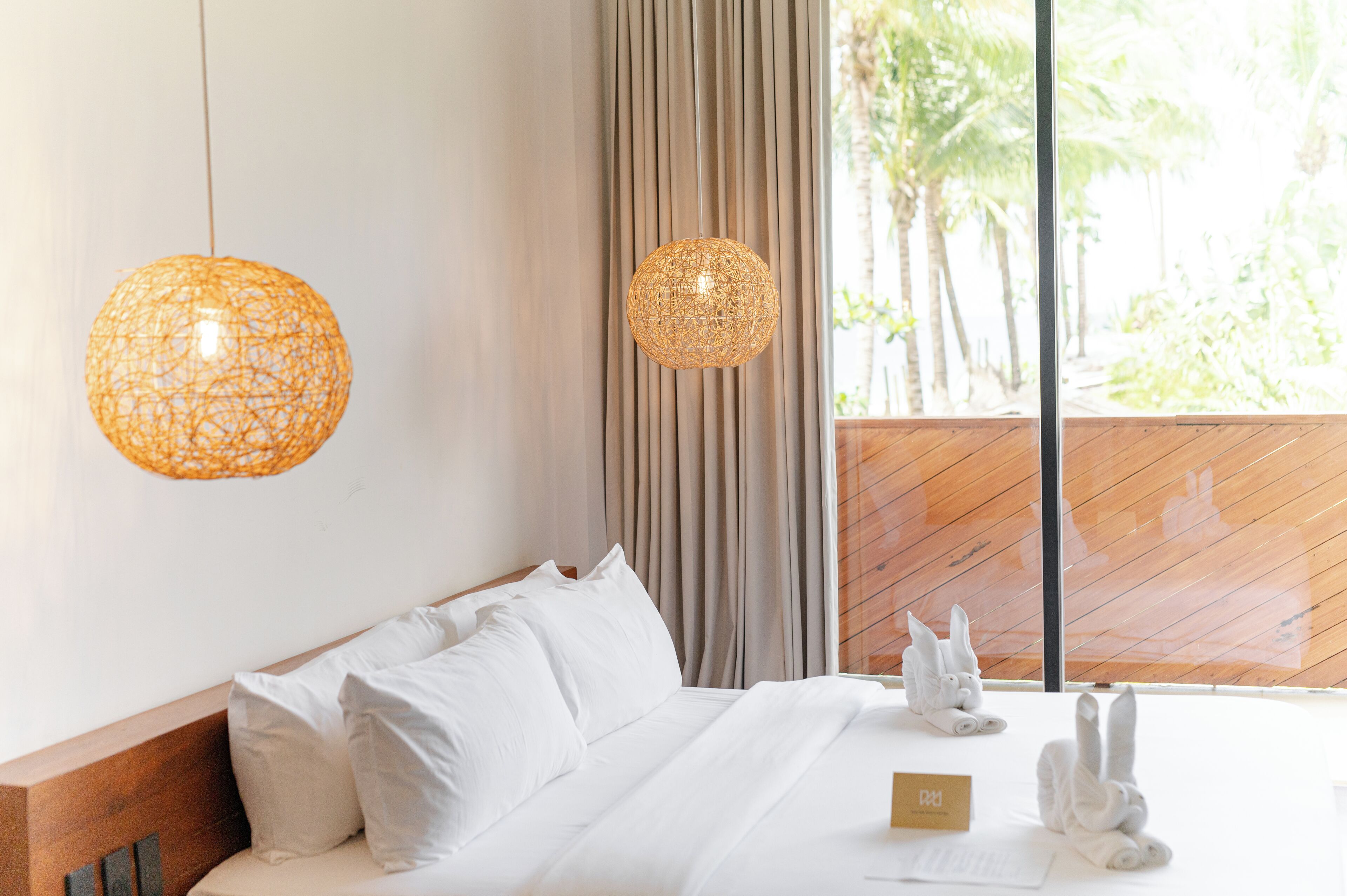 A hotel room with a bed, two hanging lamps, a window with a view of palm trees, and a wooden headboard.