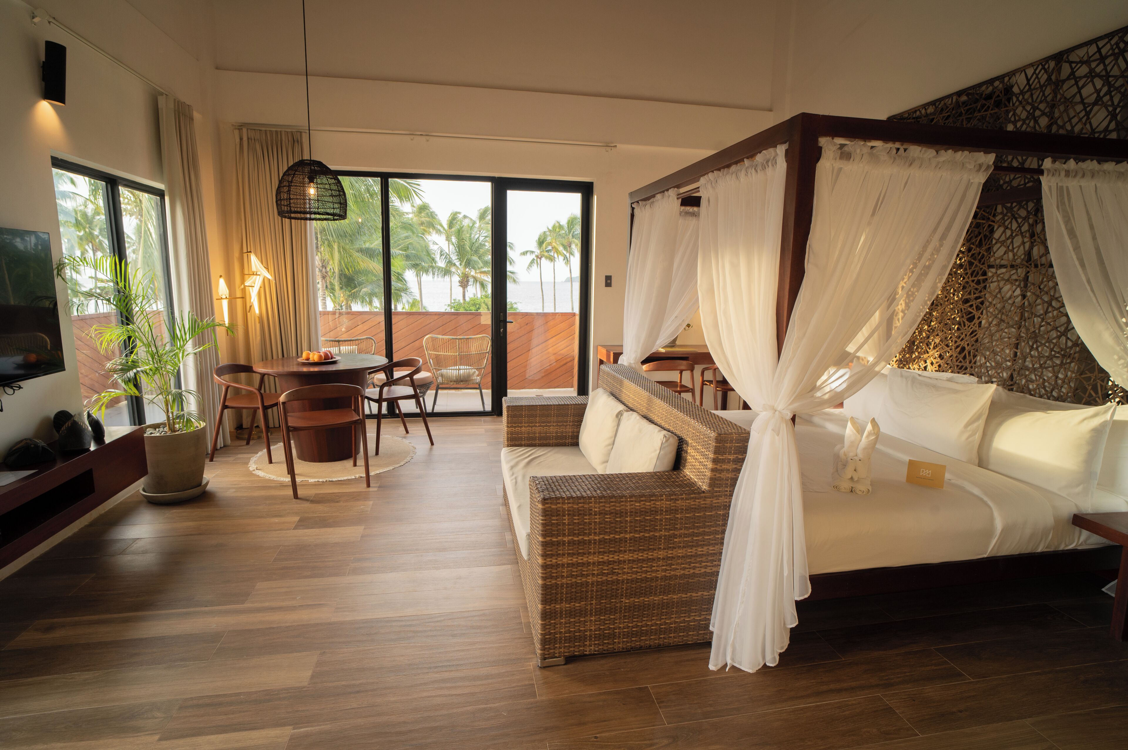 Une chambre à coucher avec un lit à baldaquin, un coin repas avec une table et des chaises, et une grande fenêtre donnant sur des palmiers.