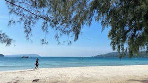 A beach with clear water, a person walking, and a boat in the distance. -  Beach  just a few steps away, offering easy access to the island’s attractions. (Preah Sihanouk)