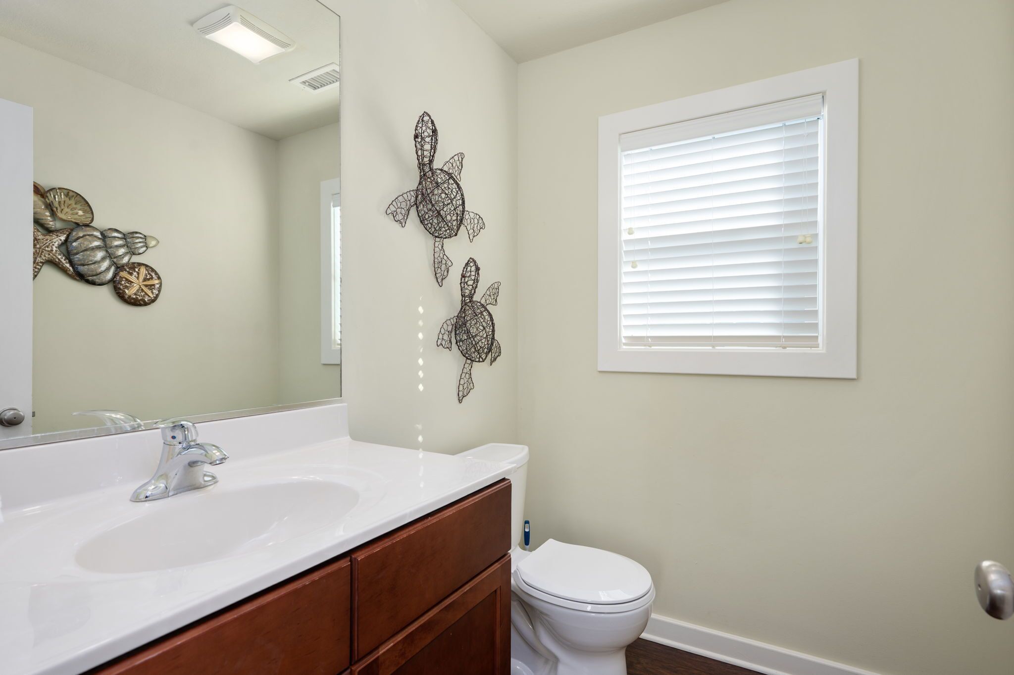 Une salle de bain avec un lavabo blanc, un meuble-lavabo en bois et des toilettes blanches. On y trouve également des éléments décoratifs sur les murs et une fenêtre avec des stores.