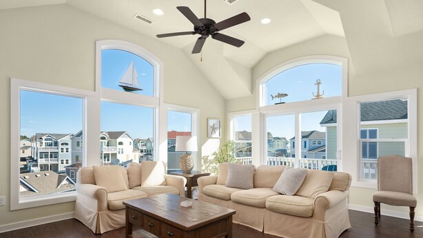 A spacious living room with a high ceiling, large windows offering a view of neighboring houses, a ceiling fan, and a wooden coffee table. - Rhapsody in Blue: Oceanside, Hot Tub, Foosball Table, Community Amenities (Corolla)
